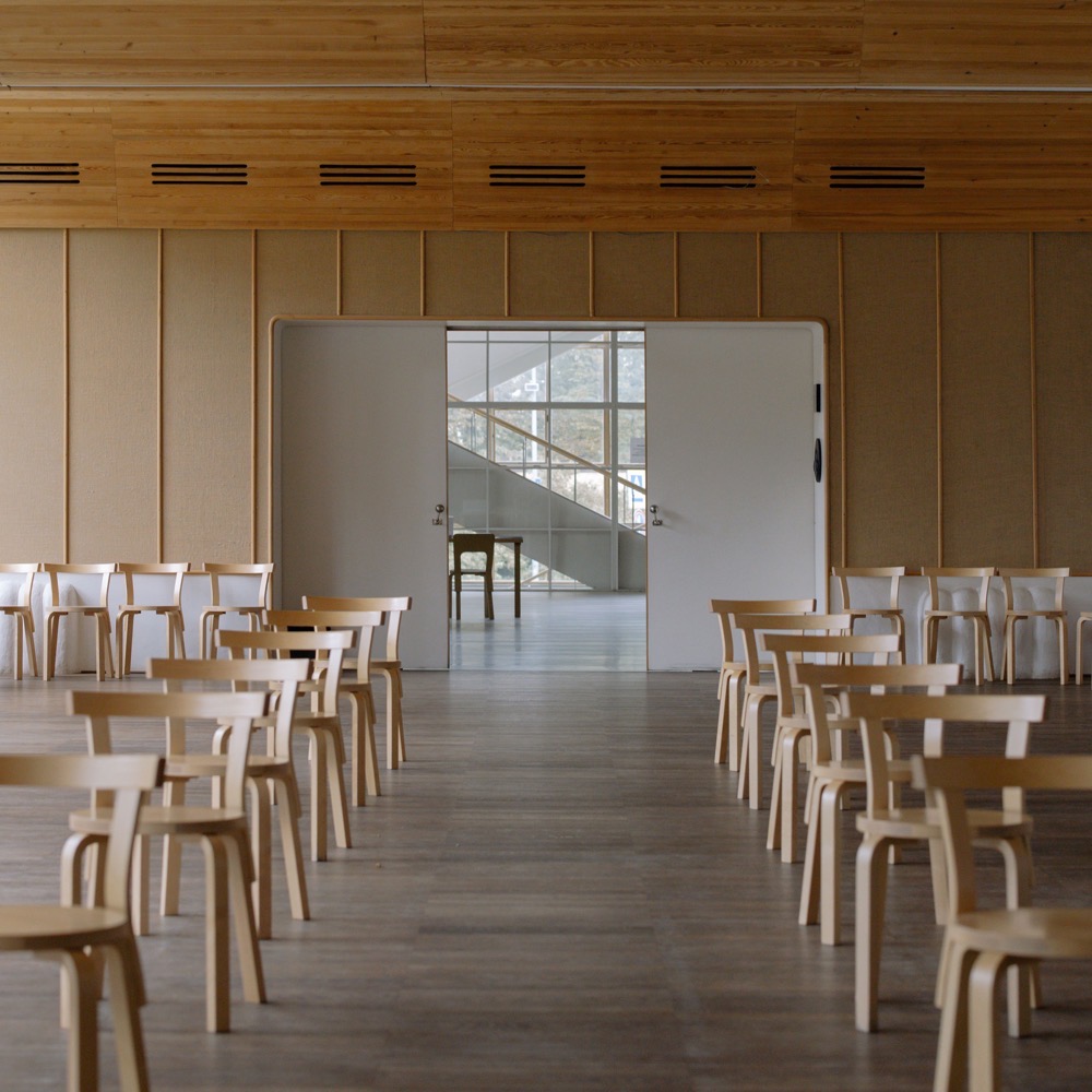 An empty, wood-panelled community hall, with rows of light wooden chairs facing a central aisle and open double doors leading to a stairwell beyond.